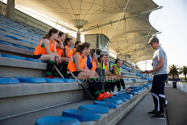 Side view of a group of female Caucasian field hockey players preparing before a game, sitting on a stand, holding hockey sticks, with their Caucasian male field hockey coach standing in front of them, talking about game plan on a sunny day — Stock Photo
