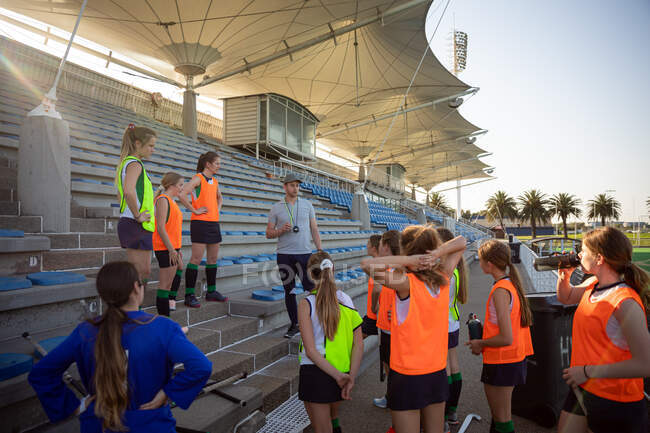 Side view of a group of female Caucasian field hockey players and their Caucasian male coach training before a game, standing on a stand, resting, listening to their coach, on a sunny day — Stock Photo