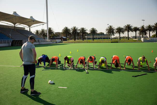 Front view of a group of female Caucasian field hockey players and their Caucasian male field hockey coach, training before a game, working out on a field hockey pitch, doing strength exercises, on a sunny day — Stock Photo