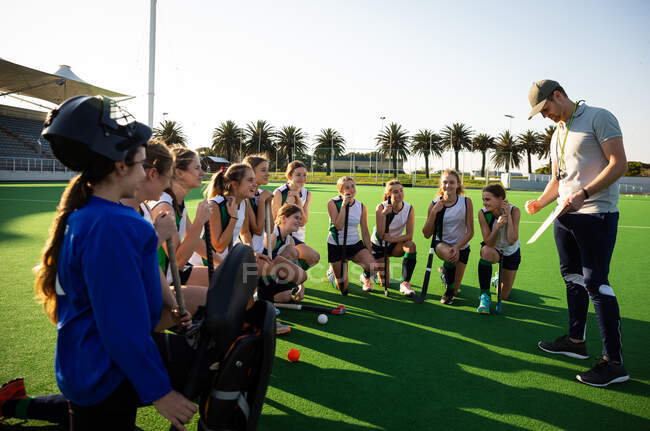 Side view of a group of female Caucasian field hockey players preparing before a game, kneeling in front of their Caucasian male coach, on a hockey pitch, who gives them game plan, on a sunny day — Stock Photo