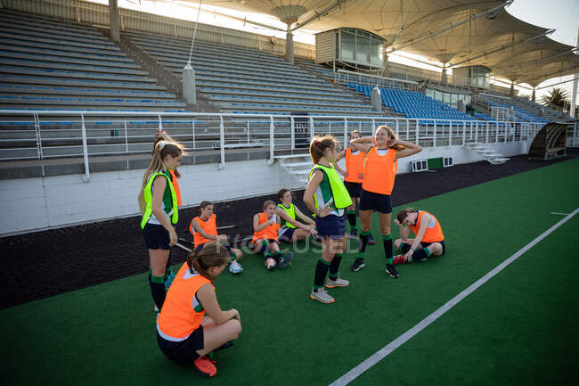 Side view of a group of female Caucasian field hockey players training before a game, standing on a hockey pitch, resting, on a sunny day — Stock Photo