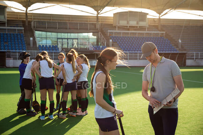 Side view of a group of female Caucasian field hockey players, preparing before a game, huddling, with one player talking to her Caucasian male coach in the foreground, on a sunny day — Stock Photo