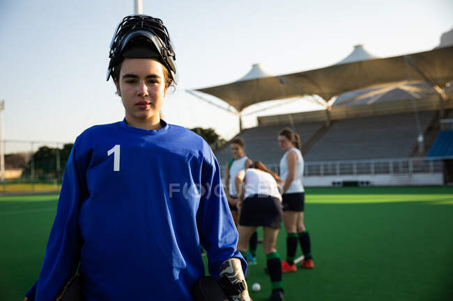 Porträt einer selbstbewussten kaukasischen Feldhockeytorhüterin, die vor einem Spiel trainiert, auf einem Hockeyfeld steht, in die Kamera blickt, einen Eishockeyhelm trägt und an einem sonnigen Tag ihre Teamkolleginnen im Hintergrund stehen — Stockfoto
