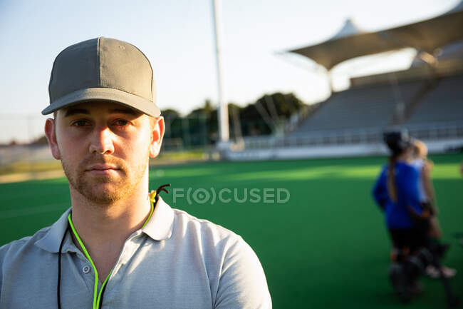 Portrait of a Caucasian male field hockey coach, preparing his team for a game, standing on a hockey pitch, looking at camera, with his team standing in the background on a sunny day — Stock Photo