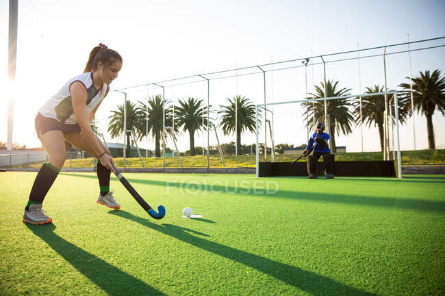 Side view of a Caucasian female field hockey player, during a field hockey game, executing a penalty kick, with a goal protected by a goalkeeper in the background, on a sunny day — Stock Photo