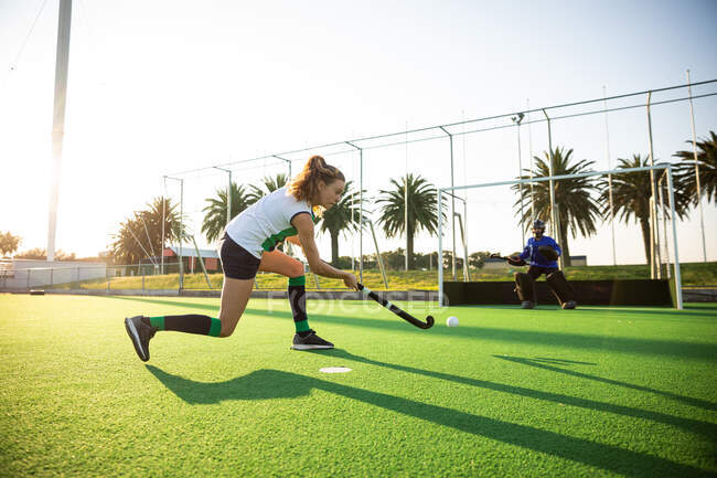 Side view of a Caucasian female field hockey player, during a field hockey game, executing a penalty kick, with a goal protected by a goalkeeper in the background, on a sunny day — Stock Photo
