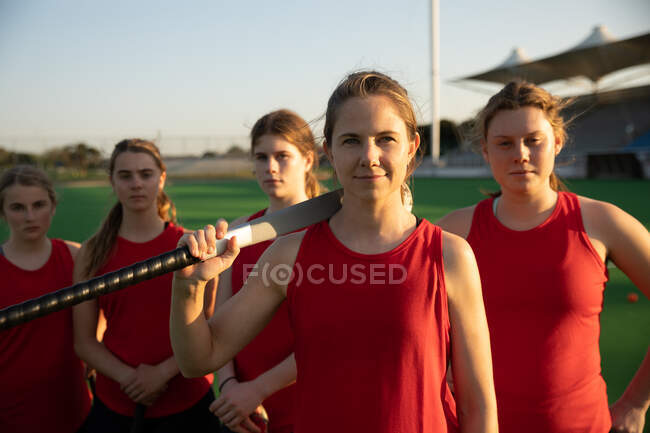 Portrait of a Caucasian female field hockey players, training before a game, standing on a hockey pitch with a hockey stick on her shoulder, looking at camera, with her teammates standing in a row behind her on a sunny day — Stock Photo