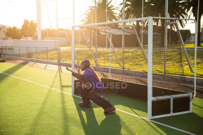 Side view of a Caucasian female field hockey goalkeeper, during a field hockey game, protecting a goal from a ball, on a sunny day — Stock Photo