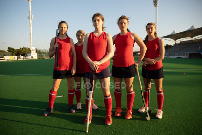Front view of a Caucasian female field hockey players, training before a game, standing on a hockey pitch, holding hockey sticks, looking at camera, on a sunny day — Stock Photo
