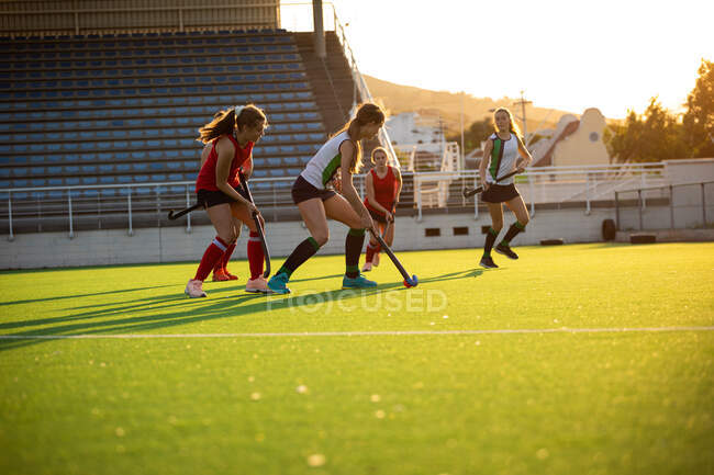 Вид сбоку кавказской хоккеистки на траве, во время хоккейной игры, бег с мячом, с хоккейной клюшкой, с ее товарищами по команде и оппонентами на заднем плане, в солнечный день — стоковое фото