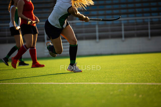 Side view low section of female field hockey player, during a field hockey game, shooting a ball, holding a hockey stick, with her teammates and opponents in the background, on a sunny day — Stock Photo