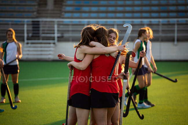 Side view of a team of female Caucasian field hockey players, during a field hockey game, celebrating a goal, with disappointed opponents standing in the background, on a sunny day — Stock Photo
