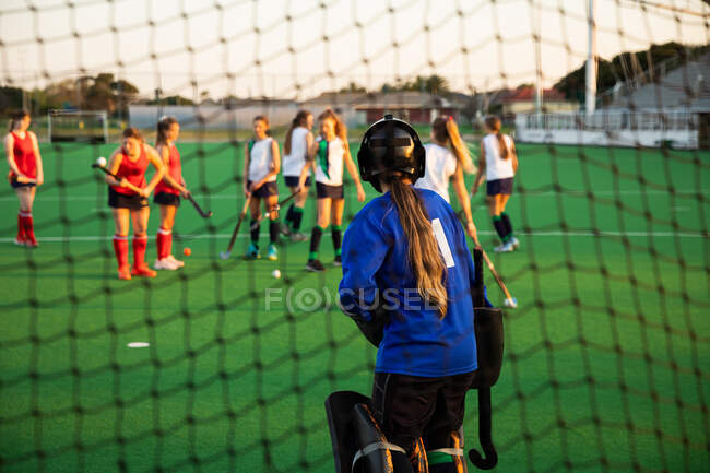 Rear view of a Caucasian female field hockey goalkeeper, during a field hockey game, standing in a goal, preparing to defend a penalty kick, with her teammates and opponents in the background, on a sunny day — Stock Photo