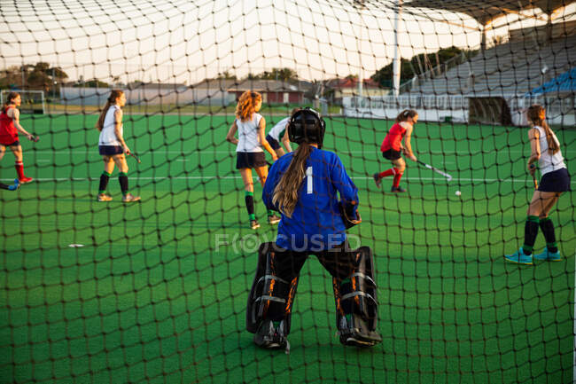 Rear view of a Caucasian female field hockey goalkeeper, during a field hockey game, standing in a goal, preparing to defend a shot, with her teammates and opponents fighting for a ball in the background, on a sunny day — Stock Photo