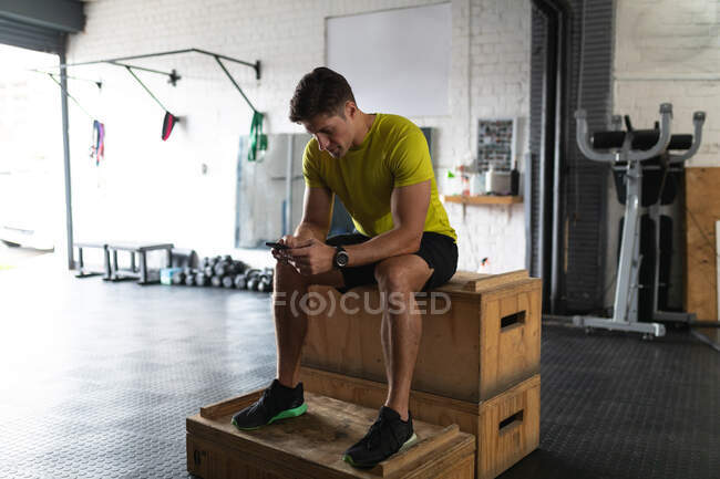Вид спереди спортсмена в спортивной одежде, тренирующегося в тренажерном зале, отдыхающего от тренировок сидящего на коробке и пользующегося смартфоном — стоковое фото