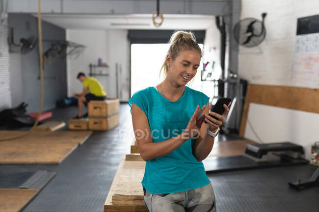 Vista frontal de una atlética mujer caucásica con ropa deportiva cruzando el entrenamiento en un gimnasio, tomando un descanso del entrenamiento apoyado en una caja, usando un teléfono inteligente y sonriendo, con un colega de gimnasio masculino sentado en el fondo - foto de stock