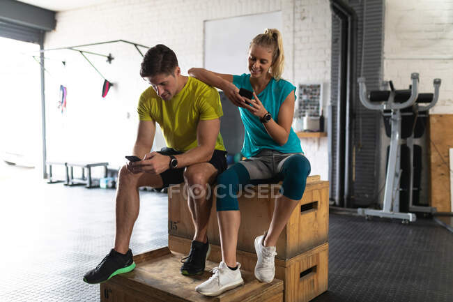 Front view of an athletic Caucasian man and woman wearing sports clothes cross training at a gym, taking a break from training sitting on a box and using their smartphones, the woman leaning on the man — Stock Photo