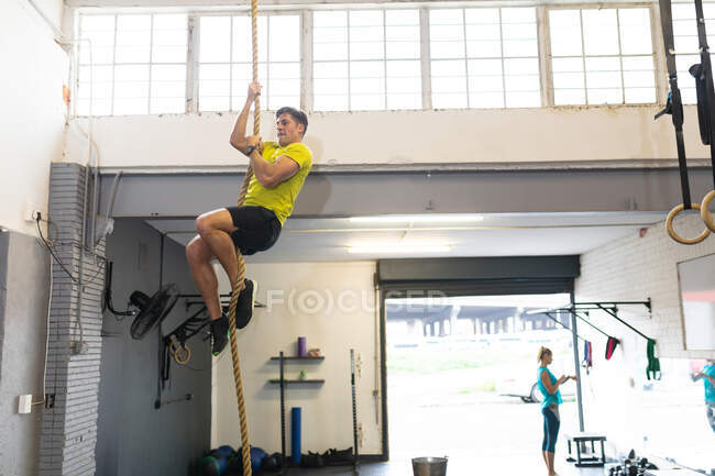 Vista lateral de um homem atlético caucasiano vestindo roupas esportivas cross training em um ginásio, escalando em uma corda, com uma mulher de sportswear em pé no fundo — Fotografia de Stock
