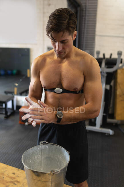 Front view of a shirtless athletic Caucasian man wearing a chest strap heart rate monitor cross training at a gym, rubbing his hands with chalk in preparation for weight training — Stock Photo
