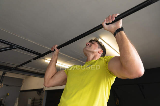 Vista lateral de cerca de un atlético hombre caucásico usando ropa deportiva entrenamiento cruzado en un gimnasio, haciendo flexiones de mentón aferrándose a un bar - foto de stock