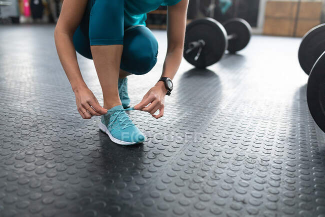 Front view low section of an athletic woman wearing sports clothes and sports shoes cross training at a gym, kneeling and tying her shoelaces before weight training with barbells, on the floor beside her — Stock Photo