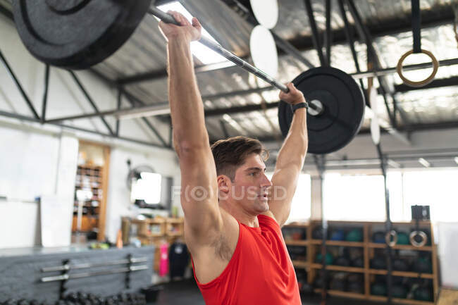 Vista lateral de un atlético hombre caucásico que usa ropa deportiva, entrenamiento cruzado en un gimnasio, entrenamiento de pie y de peso con pesas, levantando los pesos por encima de su cabeza - foto de stock