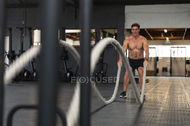 Frontansicht eines hemdlosen, athletischen kaukasischen Mannes in Sportkleidung beim Crosstraining in einem Fitnessstudio, der mit Kampfseilen trainiert — Stockfoto