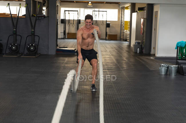 Vue de face d'un homme blanc, athlétique et torse nu, portant des vêtements de sport, s'entraînant avec des cordes de combat. — Photo de stock