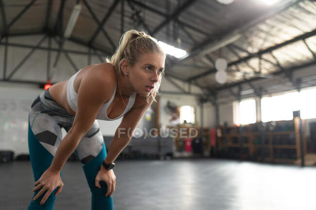 Side view of an athletic Caucasian woman wearing sports clothes cross training at a gym, standing and resting between exercises, leaning forward with her hands on her knees, looking ahead — Stock Photo