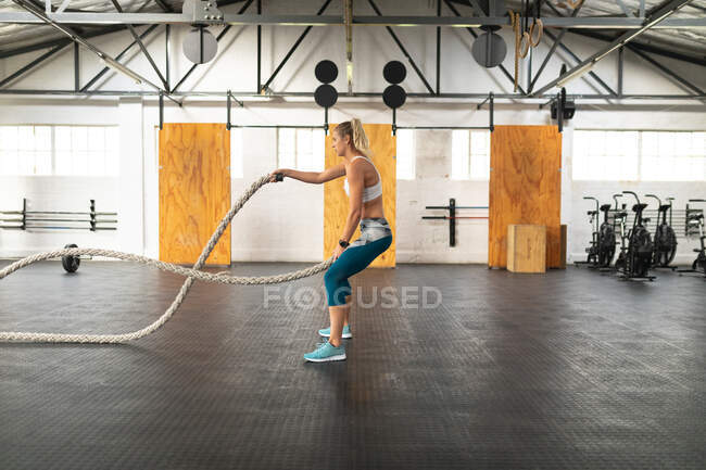 Vista lateral de uma mulher branca atlética vestindo roupas esportivas treinamento cruzado em um ginásio, trabalhando com cordas de batalha, balançando os braços com uma corda em cada mão — Fotografia de Stock