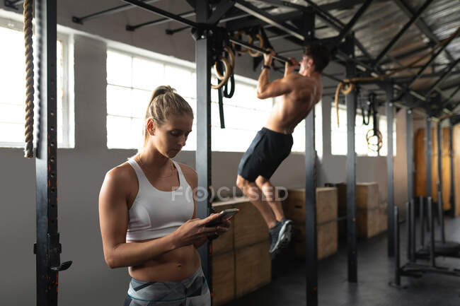 Side view of an athletic Caucasian woman wearing sports clothes cross training at a gym, taking a break from training to use a smartphone, with a shirtless athletic Caucasian man doing pull ups hanging from a bar in the background — Stock Photo