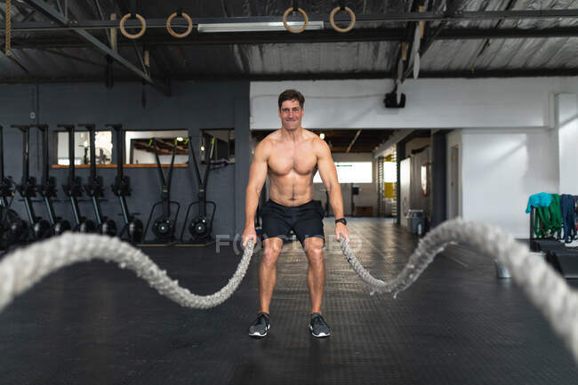 Frontansicht eines hemdlosen, athletischen kaukasischen Mannes in Sportkleidung beim Crosstraining in einem Fitnessstudio, der mit Kampfseilen trainiert — Stockfoto