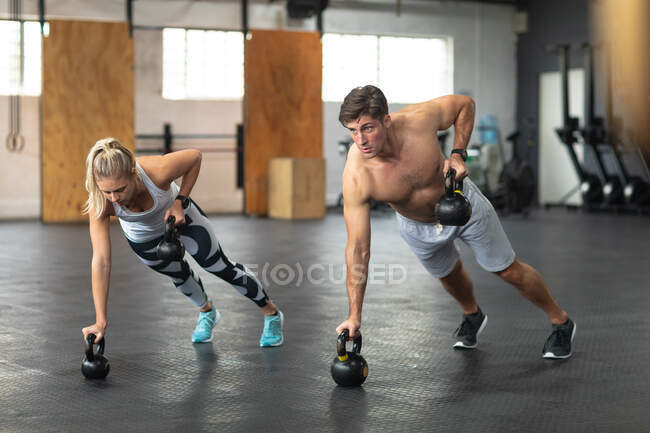 Front view of a shirtless athletic Caucasian man and woman wearing sports clothes cross training at a gym, weight training with kettlebells, leaning on one arm and lifting kettlebells with the other — Stock Photo