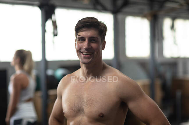 Portrait d'un homme blanc athlétique torse nu s'entraînant dans une salle de gym, prenant une pause de l'entraînement, souriant et regardant droit devant la caméra, avec une femme portant des vêtements de sport s'entraînant en arrière-plan — Photo de stock