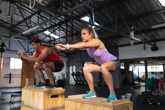 Seitenansicht eines athletischen kaukasischen Mannes und einer Frau in Sportkleidung beim Crosstraining in einem Fitnessstudio, die auf Kisten mit gebeugten Knien und ausgestreckten Armen vor sich stehen und geradeaus schauen — Stockfoto