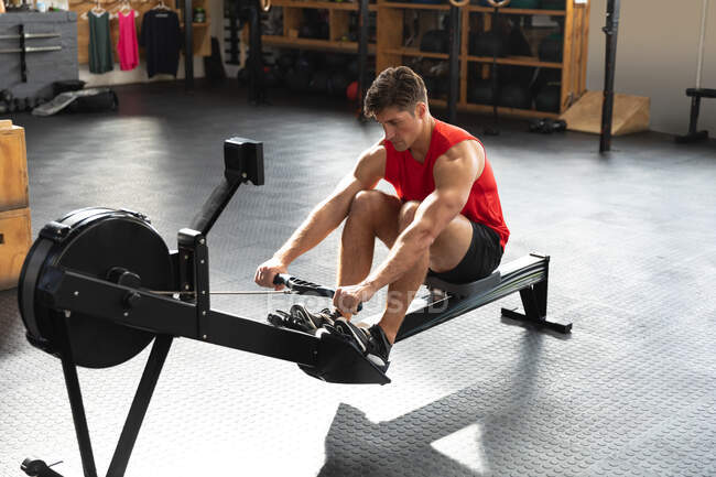 Hochwinkelaufnahme eines athletischen kaukasischen Mannes in Sportkleidung beim Crosstraining in einem Fitnessstudio, der mit erhobenen Knien sitzt und auf einem Rudergerät trainiert — Stockfoto