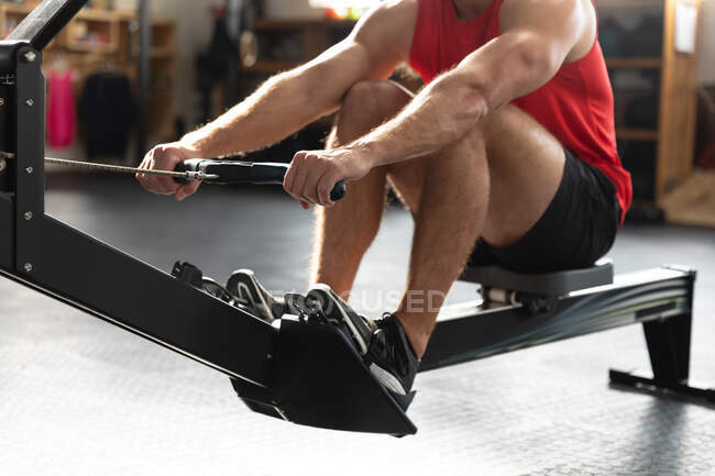 Vista frontal de la sección central de un atleta que usa ropa deportiva, entrenamiento cruzado en un gimnasio, sentado con las rodillas arriba, haciendo ejercicio en un remo. - foto de stock