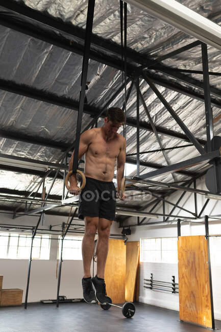 Frontansicht eines hemdlosen athletischen kaukasischen Mannes beim Crosstraining in einem Fitnessstudio, der sich an Turnringen hochschiebt und sein Körpergewicht hebt — Stockfoto