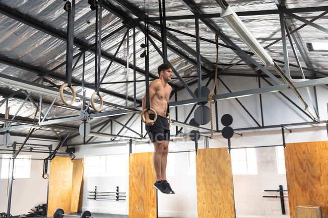 Вид сбоку на спортсмена без рубашки, тренирующегося в спортзале, подталкивающего себя на гимнастические кольца, поднимающего вес тела — стоковое фото