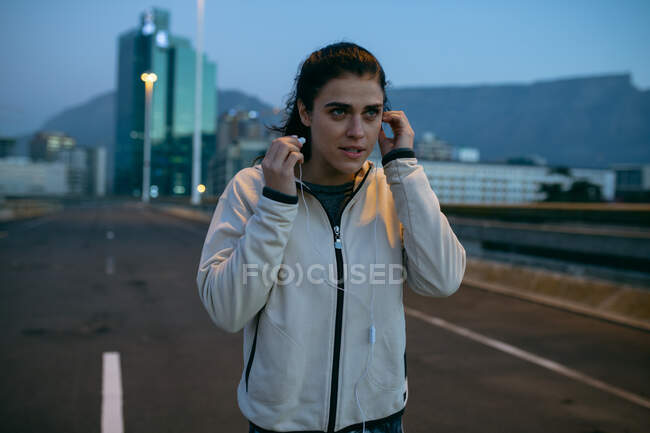 Front view of a fit Caucasian woman with long dark hair wearing sportswear exercising outdoors in the city in the evening, running putting her earphones on, with urban buildings in the background. — Stock Photo