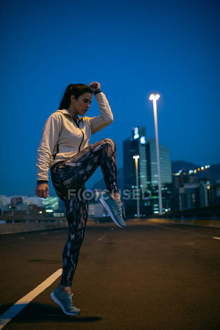 Low angle side view of a fit Caucasian woman with long dark hair wearing sportswear exercising outdoors in the city in the evening, warming up, stretching her leg with urban buildings in the background. — Stock Photo