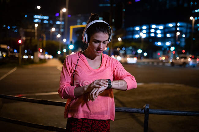 Front view of a fit Caucasian woman with long dark hair wearing sportswear exercising outdoors in the city in the evening, standing checking her smartwatch with headphones on with urban buildings in the background. — Stock Photo