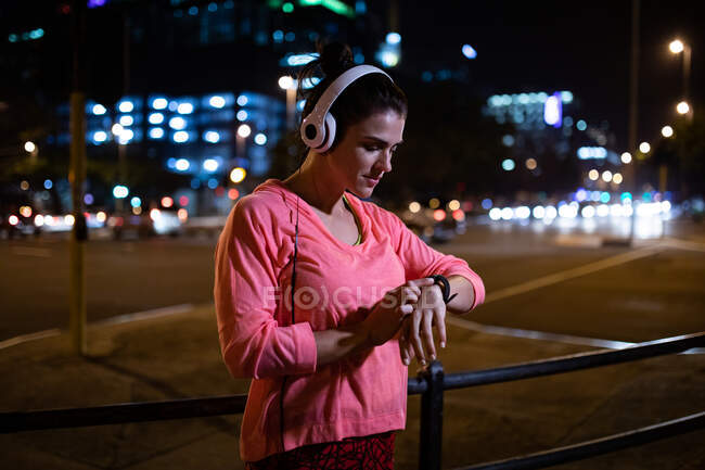 Бічний вид на кавказьку жінку з довгим темним волоссям у спортивній манері, яка вправляється на відкритому повітрі у місті ввечері, стоячи, перевіряючи свій смартлет з навушниками на задньому плані з міськими будівлями.. — стокове фото