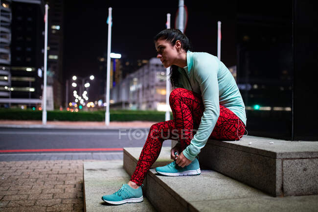 Seitenansicht einer fitten kaukasischen Frau mit langen dunklen Haaren, die abends im Freien in der Stadt Sport treibt und eine Pause von ihrem Training macht, sitzend auf einer Treppe, die Schnürsenkel bindet, mit städtischen Gebäuden im Hintergrund.. — Stockfoto