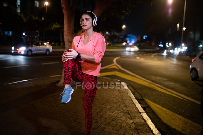 Vorderansicht einer fitten kaukasischen Frau mit langen dunklen Haaren, die abends im Freien in der Stadt Sport treibt, sich aufwärmt, sich mit Kopfhörern dehnt und im Hintergrund städtische Gebäude sieht. — Stockfoto