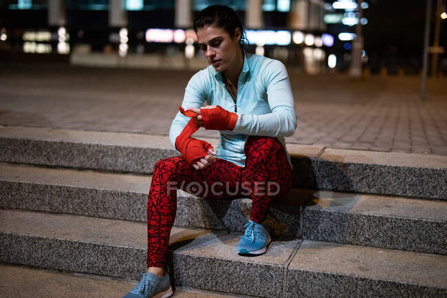 Vorderansicht einer durchtrainierten kaukasischen Frau mit langen dunklen Haaren, die abends im Freien in der Stadt Sport treibt, auf Stufen sitzt und ihre Hände mit Boxband umwickelt, im Hintergrund Gebäude — Stockfoto