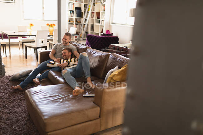 Vista frontal de pareja masculina caucásica relajándose en casa, sentada en un sofá, abrazando, interactuando y sonriendo — Stock Photo