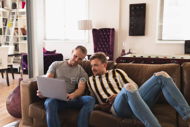 Vorderansicht eines kaukasischen männlichen Paares, das es sich zu Hause gemütlich macht, auf einem Sofa sitzt und mit einem Laptop interagiert — Stockfoto