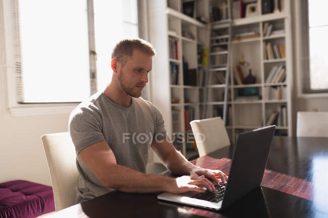 Side view of a young Caucasian man spending time at home, sitting by the desk and using his laptop computer. — Stock Photo