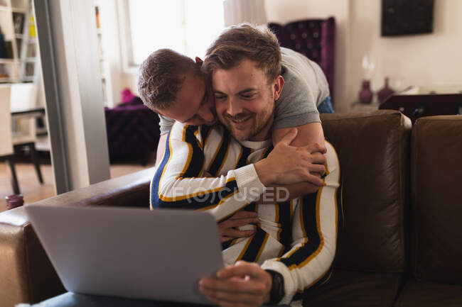 Вид спереди на белых мужчин, расслабляющихся дома, сидящих на диване, обнимающихся, взаимодействующих друг с другом с помощью ноутбука — стоковое фото